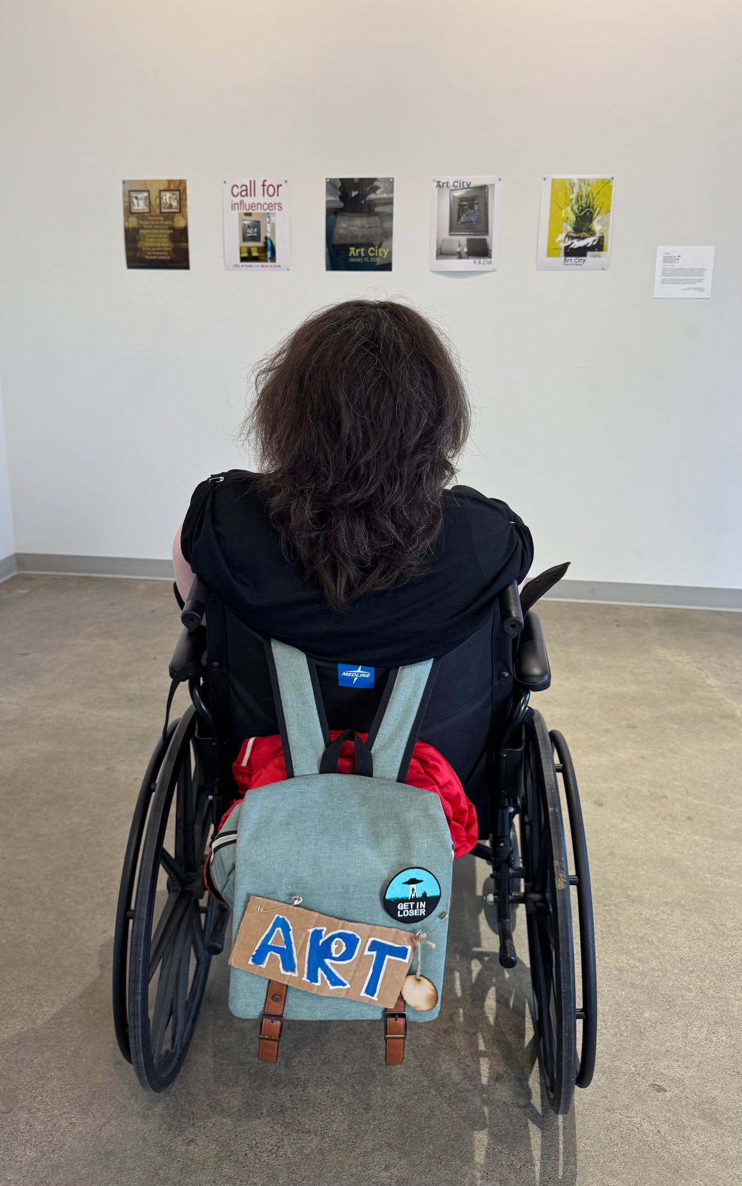 I am sitting in my wheel chair looking at the show posters. My hair is getting long and my backpack has a sign that says "art." Photo credit Ember DeBoer