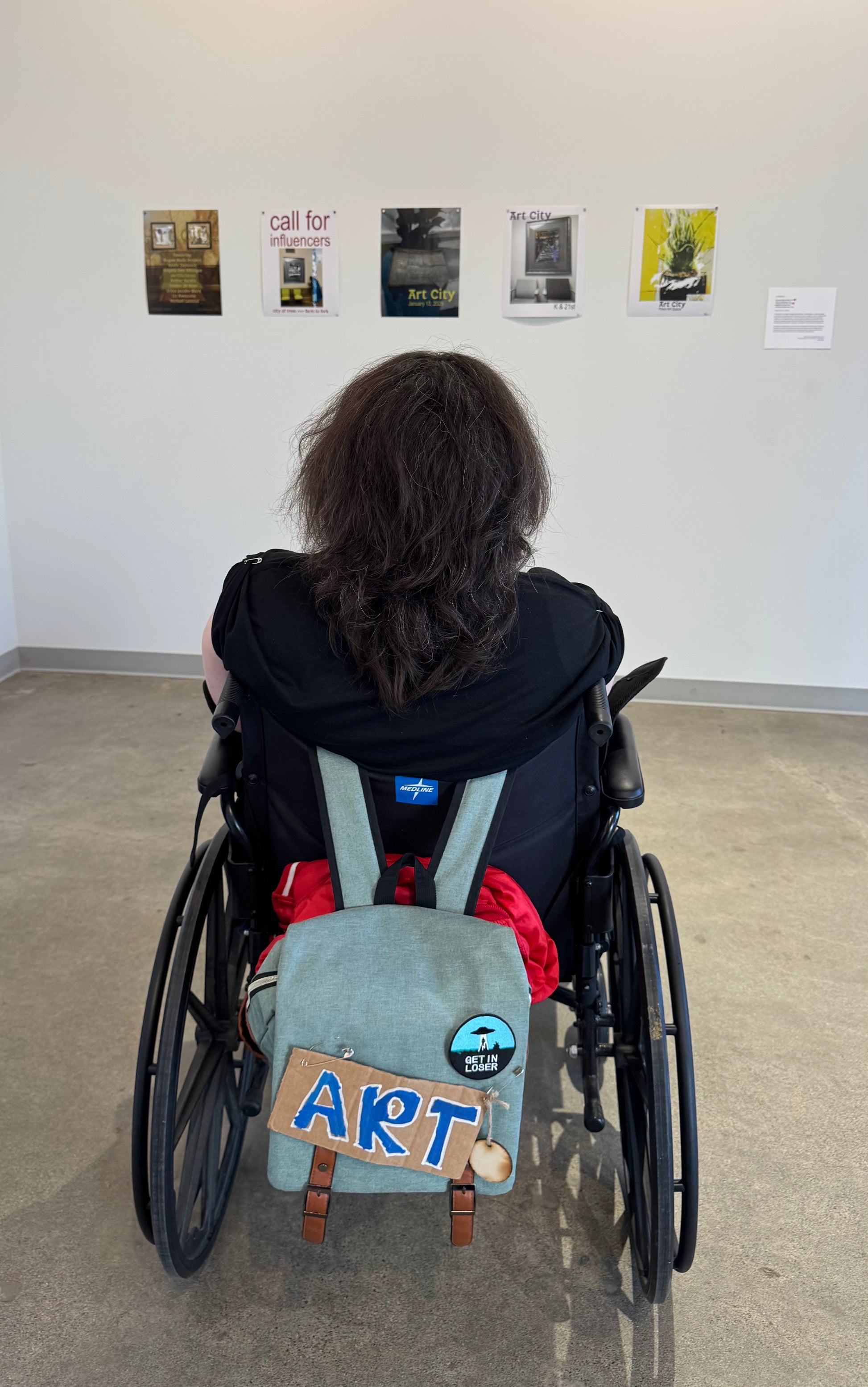 I am sitting in my wheel chair looking at the show posters. My hair is getting long and my backpack has a sign that says "art." Photo credit Ember DeBoer